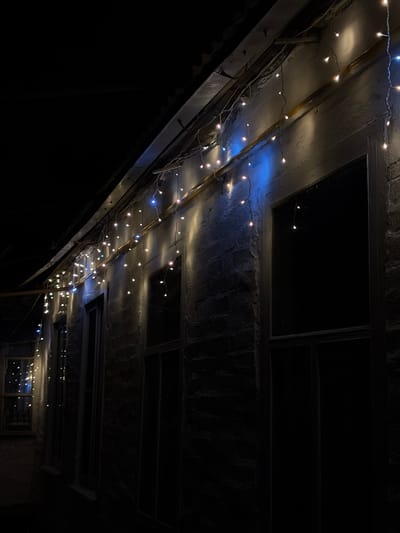 The media file displays a low-angle shot of a ceiling adorned with strings of small, white and blue fairy lights. These lights are draped across what appears to be an interior or covered outdoor space, with a rough, textured ceiling that has recessed rectangular panels. The overall impression is dark and moody, with the fairy lights providing the primary illumination. Some of the lights have a warm, incandescent glow, while others emit a cooler, blue light. The ceiling structure suggests it could be an attic, a covered walkway, or a similar architectural feature with exposed beams and panels. The darkness implies it is either night or a very dimly lit indoor environment. There is no text visible in the image. The scene is devoid of people, focusing entirely on the decorative lighting and the architectural elements of the ceiling. The mood evoked is somewhat festive due to the lights, but also mysterious and perhaps a bit desolate due to the darkness and the starkness of the setting.