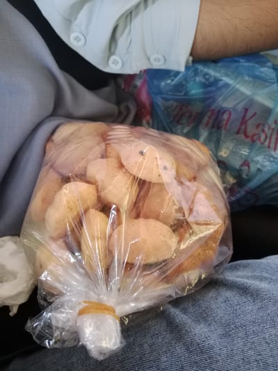 A plastic bag of small, rounded baked goods rests on a pair of denim jeans. The bag is sealed with a yellow rubber band. In the background are other clothing items and a blue plastic bag printed with the words "Terima Kasih". The location is Kruet Teumpeun, Indonesia, as provided. There are no people fully visible, only an arm and clothing.