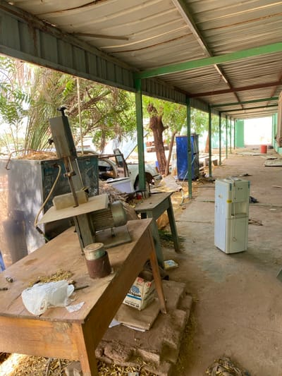 The image shows a scene under a covered outdoor area in Khartoum, Sudan.  The coordinates 15.552, 32.57 correspond to a location in Khartoum, Sudan. The scene appears to be a workshop or storage area with a metalworking machine, a wooden table, and a water cooler. The machine is a band saw with a motor and a metal cutting blade. The table is made of wood and has a metal frame. The water cooler is white and appears to be in working condition. There is debris on the ground, consisting of dried leaves, and other miscellaneous items.  The scene is dusty and appears to be in disuse. No people are present.  The area is enclosed by a metal roof supported by metal beams. The roof is green in color and has a rusty appearance.  The environment appears dry and arid.  The ground is rough and uneven. The scene is lit by natural light.  The surrounding area is not visible.