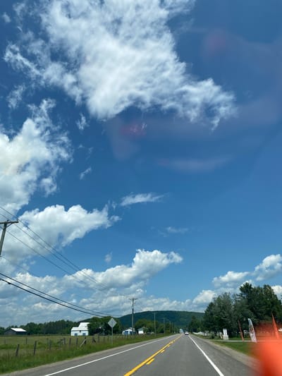 This is a vertical, medium shot of a rural road scene in Saint-André-Avellin, Canada, under a bright blue sky dotted with fluffy white clouds. The road, paved and divided by yellow lines, stretches into the distance, flanked by a grassy shoulder and a white line. To the left of the road, a wooden fence encloses a green field containing a dark barn and a white building with blue accents. Power lines run diagonally across the frame from left to right, supported by utility poles. In the background, a line of trees and rolling green hills form the horizon. On the right side of the road, several trees stand, and further down, a white fence and some orange flags are visible. The overall impression is one of a sunny, clear day in a tranquil countryside setting.