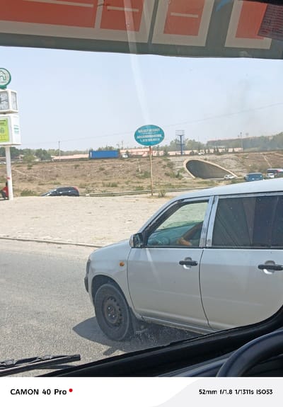 The image is taken from inside a vehicle, looking out at a roadside scene. A white car is prominent in the foreground, while in the background there is a highway with a tunnel, other vehicles, and a sign for "Galaxy Resort" with a phone number. The weather appears sunny. The camera details are visible at the bottom of the image.