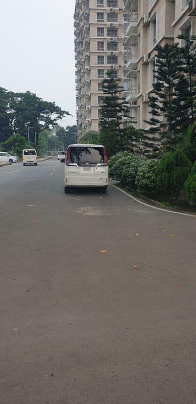 The image depicts a street scene in Chor Sahapur, Bangladesh, with a focus on a white minivan parked on the right side of the asphalt road. The minivan, identified as a Toyota Noah based on its visible badging, has its rear facing the camera. The license plate is partially obscured but appears to have Bengali script and numbers. The road itself is paved and has yellow lane markings. In the background, a multi-story building with cream-colored facades and numerous balconies dominates the right side of the frame. Lush green trees and manicured bushes line the side of the road and the building. A white car is visible further down the road on the left, and another white minivan is seen in the distance, also traveling on the road. The sky is overcast and appears light gray, suggesting a cloudy day. The overall environment appears to be a well-maintained urban or suburban area with a mix of residential and possibly commercial structures, as indicated by the tall building. There are no people visible in the immediate vicinity of the minivan.