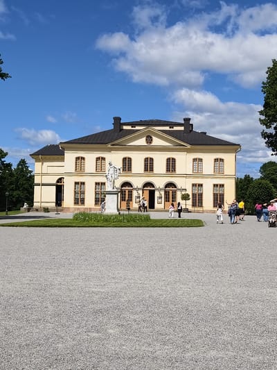 The image depicts the Drottningholm Palace, located in Ekerö kommun, Sweden. The palace is a grand yellow building with a dark roof, featuring numerous windows with wooden frames. In front of the palace, a white marble statue stands on a pedestal, surrounded by a manicured lawn and flower beds. Several people are visible on the gravel path leading to the palace, some appearing to be tourists. The sky is blue with scattered white clouds, and trees frame the scene on either side, suggesting a pleasant outdoor environment. The foreground is dominated by a wide expanse of grey gravel.