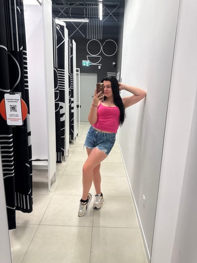 The media file is a full-body mirror selfie of a young woman in what appears to be a retail fitting room or display area. She is standing and taking the photo with her phone. She has long, dark hair and is wearing a bright pink tank top and distressed denim shorts. She is also wearing white sneakers with grey and orange accents. The woman's pose is relaxed, with one arm behind her head and the other holding her phone.
The background shows a clean, modern interior with light-colored tiled floors. To the left of the woman, there is a wall with large black and white graphic art that appears to be text or a logo. Further to the left, there are tall, white display shelves with black fabric draped over them, featuring repeating white geometric patterns. A QR code on an orange circular sticker is visible on one of the shelves, along with some text. Visible text includes "TЪРСИТЕ НЕЩО РАЗЛИЧНО?" and "НАМЕРЕТЕ ОЩЕ ПО-ВЕЧЕ МОДЕЛИ В ПРИЛОЖЕНИЕТО НА SINIZATI". This suggests the location is a store in Skutare, Bulgaria. The lighting appears to be bright and even, suggesting it's daytime.