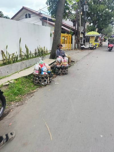 A man sits on the side of a road next to two baskets of colorful packaged goods. A tree trunk serves as his backrest. A white wall borders the road with tall plants. A food cart and passing vehicles are visible further down the road. A sandal-clad foot is visible in the bottom left corner of the frame. The road is gray and stretches into the distance.