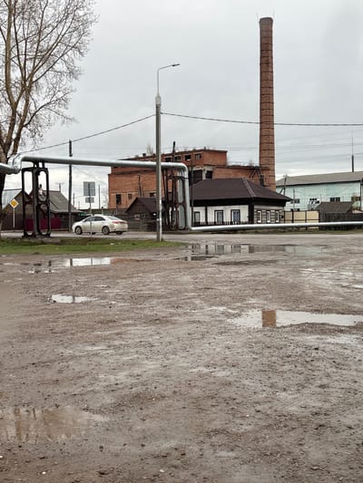 The image depicts a wet, muddy street scene in Ust-Ordynsky, Russia. A tall brick chimney stands prominently near several buildings, one with dark brown trim. A white car is parked near a grassy patch and a road sign. Overhead pipes cross the foreground. The sky is overcast, and a bare tree is visible in the lower left.
