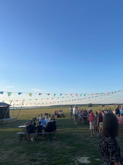 The image depicts an outdoor gathering at sunset near Am Salzhaff, Germany. A clear blue sky with sparse clouds forms the backdrop, with several strings of colorful pennant flags strung across the scene, some illuminated by string lights. In the foreground, the grass is mowed, with patches of dry soil.
To the left, a small stage structure is set up, with a person on it holding a guitar, suggesting live music. In front of the stage, a few people are seated at picnic tables. Further back, many people are scattered throughout the scene, standing and sitting around various picnic tables arranged on the grassy field. Some individuals appear to be eating or conversing. The middle ground shows a body of water, likely the Salzhaff, with a treeline visible on the horizon. The overall atmosphere suggests a relaxed, communal event, possibly a festival or a summer celebration.