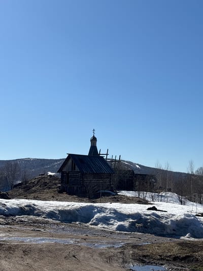 The image shows a wooden church standing near a snow-covered slope under a clear blue sky. The church has a distinct steeple with a cross on top. Part of a dirt road runs alongside the snow. The location is the Gubakha region in Russia.