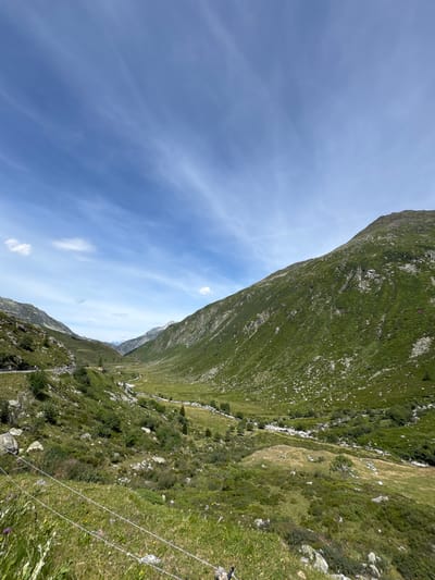 This is a daytime, eye-level, wide-angle photograph of a lush green mountain valley in Hospental, Switzerland. The sky is a clear blue with wispy white clouds. The valley is flanked by steep, grassy mountainsides dotted with rocks and sparse trees. A winding road is visible on the left side of the valley, and a small stream or river meanders through the valley floor. In the foreground, a wire fence runs horizontally across the image. There are no people or animals immediately visible in the scene. The overall impression is one of natural beauty and rugged terrain.