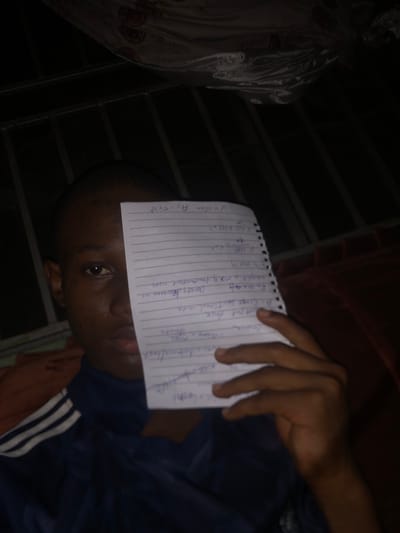 A person is holding a notebook with handwritten notes in front of their face. The notebook contains formulas and notes related to physics or engineering, including terms like "Cross Sectional area," "Density decrease," "weight = mxg," "V=10mm," and "A=1.5x18". The person is wearing a blue shirt with white stripes on the shoulder. The background is dark, but hints of a window and bedding are visible. The location is not specified and can not be identified.