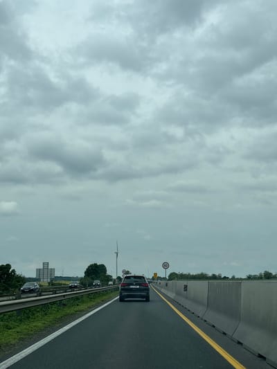 The image depicts a multi-lane highway in the vicinity of Ács, Hungary, under an overcast sky. Traffic is moderate, with several cars visible in the lane. A concrete barrier separates the highway from the adjacent roadside. Signage, including a speed limit sign (60), is present. In the distance, a wind turbine is visible. Vegetation is also present on the roadside.