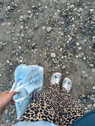 The image shows a person's legs and feet standing on a gravel path. The person is wearing a leopard print skirt and floral patterned Crocs. They are holding a white plastic bag filled with items. The ground is covered in various sizes of grey, brown, and white stones and dirt. The image appears to be taken from a high angle, looking down at the person's legs and the ground. The specific location in Abushkovo, Russia is not discernible from the image.