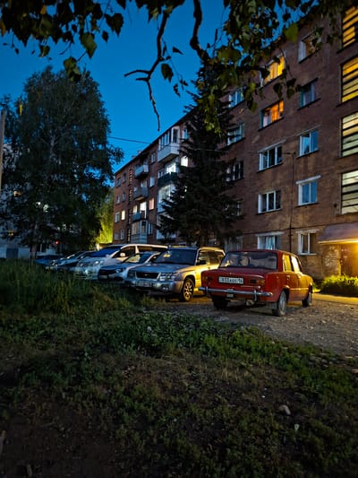 The image depicts a nighttime scene in Ridder, Kazakhstan, with a focus on a row of parked cars alongside a brick apartment building. The building's windows are illuminated, suggesting people are inside, creating a warm contrast with the dark sky. A tree with green leaves in the foreground partially obscures the view of the building.
Several cars are parked in a line, one of which is a red Soviet-era Lada with the license plate "833 AGS 16". Other vehicles are parked behind it, including what appears to be a silver SUV with the license plate "734 ZBA 16". The cars are parked on a slightly muddy, grassy verge next to a road. The overall atmosphere is one of quiet residential life at dusk or early night. The sky is a deep blue, indicative of twilight or nighttime. The lighting from the building and streetlights casts a soft glow on the scene.