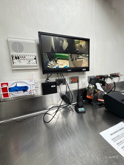 This media file captures a close-up, angled view of a security monitoring station and surrounding equipment, likely in a commercial kitchen or restaurant setting in Panama City Beach, USA. The primary focus is a flat-screen monitor displaying a multi-camera feed, showing various interior views including what appears to be a kitchen area with stacked cans and other equipment. To the left of the monitor is a white intercom or control panel with numerous buttons.
Below the monitor, a box of blue vinyl gloves is mounted in a white wire rack. To the right of the monitor, a silver metal countertop holds several electronic devices. These include a two-way radio with "KITCHEN" clearly visible on its side, a black electronic device, and a charger. Power outlets are visible on the white wall behind the equipment. A paper menu, likely from "DAVE & BUSTER'S," is also present on the countertop, with visible text listing menu items such as "MOUNTAIN OF NACHOS" and "PRETZEL DOOD." The overall scene suggests a functional, operational area, possibly during a business day, given the presence of the intercom and security setup. No people are visible in the frame. The lighting appears to be artificial, typical of an indoor environment.