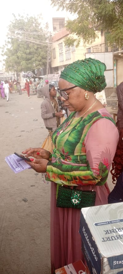 A woman with glasses wearing a green beaded headwrap, a green floral top, and a light pink dress stands outside holding a phone. She also has a green beaded purse with crystal embellishments. In the background is a street scene with people, vehicles, and buildings. A box partially visible in the foreground has "Hollandia" and "Yoghurt Drink" printed on it.