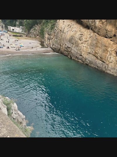 The image depicts a picturesque beach with crystal-clear turquoise water. The beach is a mix of sand and pebbles, with several sunbathers lounging on chairs. A small stream empties into the sea at one end of the beach. The beach is nestled between towering, rugged cliffs, with lush greenery clinging to their sides. In the background, white buildings with red-tiled roofs are visible, suggesting a small coastal village. The overall impression is one of tranquility and natural beauty. It's unlikely this is Norcia, Italy, as Norcia is landlocked and not a coastal town.
