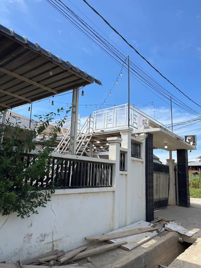 The image depicts an outdoor scene in Tanjungkarang, Indonesia, featuring a building with an attached outdoor staircase and rooftop balcony. A white wall with a black metal fence runs along the front of the property. Some construction materials, including wooden planks and white panels, are scattered near the wall and in a ditch. Overhead, power lines and decorative string lights are visible against a bright blue sky with scattered clouds. A lush green bush is positioned to the left of the wall. The building appears to be two stories high, with the staircase leading to an elevated platform. A sign with a black silhouette and text is mounted on the right side of the building's facade. The area surrounding the building seems to be a pathway or street, with a glimpse of vegetation and other buildings in the background.