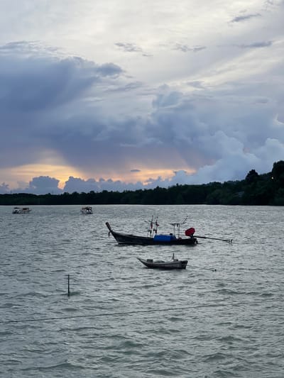 The image depicts a tranquil scene of a body of water with several boats. The water is choppy, suggesting a slight breeze. In the foreground, a long, dark boat is anchored, equipped with fishing rods. A smaller, lighter-colored boat is moored nearby. Further in the distance, two other small boats are visible. The background features a dense, dark green treeline, likely a forested hillside, with a few lights suggesting human habitation.
The sky is dramatic, with a mix of dark, heavy clouds and patches of bright sunlight breaking through, casting a warm, golden glow. This contrast suggests either a storm approaching or receding, or the sun setting. The overall mood is serene and perhaps a little dramatic due to the sky. The time of day appears to be late afternoon or early evening. There is no visible text in the image. The location is identified as Pa Khlok, Thailand, which is consistent with a coastal or lakeside setting with lush vegetation.
