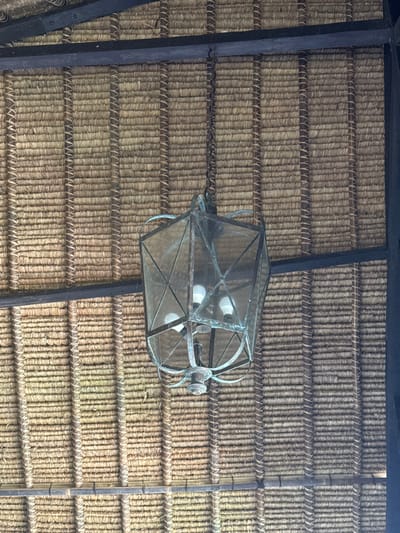 The image depicts an overhead view of a hexagonal, antique-style lantern fixture suspended from a natural fiber ceiling in Tibubiyu, Indonesia. The fixture, likely constructed of patinated metal with glass panels, contains three visible incandescent bulbs. It hangs from a dark metal chain attached to a prominent dark wooden beam that spans horizontally across the upper portion of the frame. The ceiling consists of tightly woven, light brown, straw-like material arranged in horizontal rows with darker, possibly bundled, vertical supports creating a patterned texture. Additional dark structural beams are partially visible on the right and lower sections of the image. No people or specific actions are visible within the frame.