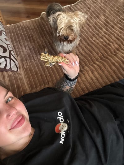 A woman lies on a brown corduroy sofa. A dog is positioned beside her, near her left hand, and she holds a gold-colored, rope-like object in her fingers. The woman has tattoos on her left arm, a nose piercing, and freckles. She is wearing a black sweatshirt with the word "Nala" visible in white print. The location is unknown and it is not clear that it is in the city of Chornomorskoe, Ukraine.