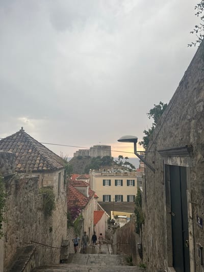 The image depicts a narrow, stone-paved street in Dubrovnik, Croatia, on an overcast day. The viewpoint is looking down the street, which slopes slightly downwards towards the sea. The architecture is consistent with the Old Town of Dubrovnik, featuring ancient stone buildings with terracotta tiled roofs.
In the foreground, the rough texture of stone walls and a tiled roof are prominent. A street lamp protrudes from a building on the right. Further down the street, several people are visible, walking and standing. They are dressed casually, suggesting a normal day rather than a specific event.
Beyond the buildings, a hazy view of the sea can be seen, with a distant fort or castle perched on a hill. The sky is heavily clouded, creating a diffused light that suggests either early morning or late afternoon. The overall mood is subdued, reflecting the overcast weather.
There is a small blue sign with white lettering on a building on the right side of the image, but the text is not legible. The scene captures the characteristic charm of Dubrovnik's historic streets.