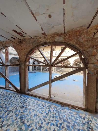 The image shows a view from the inside of a building, looking upwards. The building appears to be in a state of disrepair, with cracked and peeling plaster on the walls and a large, exposed wooden frame in the center of the image. The frame is positioned over a space with a light blue floor, which appears to be a tiled surface. The building is located in Oran, Algeria (35.703, -0.647). The floor is covered with a mosaic of small, irregular blue, white, and beige tiles. The wooden frame is constructed of weathered wooden beams and crossbeams. The view beyond the frame is obstructed by the wooden structure, but the light blue floor can be seen through the gaps in the frame.