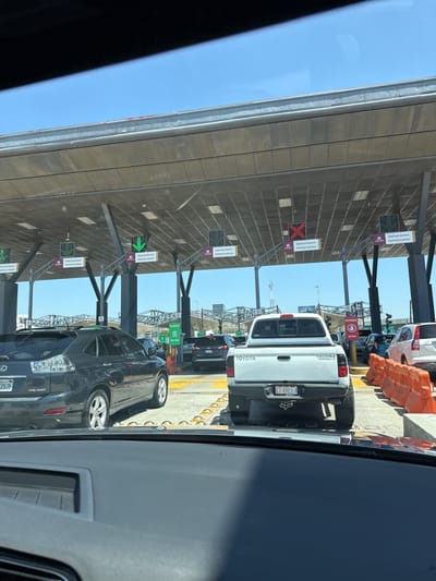 The image shows a queue of vehicles at a border crossing in Tijuana, Mexico. Overhead structures feature signs with green arrows and red Xs, indicating lane availability, along with text that reads "Nada que declarar" in Spanish and "Nothing to declare" in English. Vehicles visible include a dark SUV, a white Toyota Tacoma truck, and a white SUV. The license plate of the Toyota Tacoma is visible. Orange traffic barriers are on the right side of the image. The sky is clear and blue.