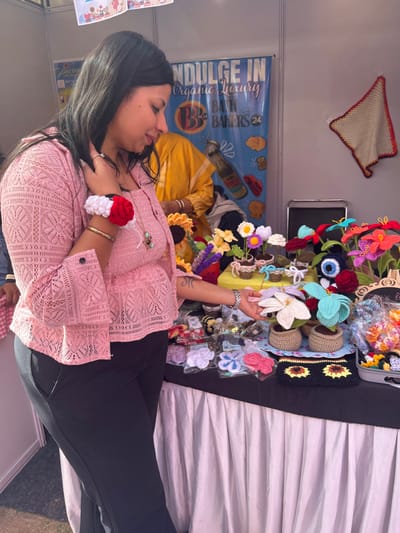 The image shows a woman in a pink lace top and black pants standing in front of a stall selling handmade crocheted flowers. The stall is located in Sector 17, Chandigarh, India (latitude: 30.764, longitude: 76.798). The woman is looking at the crocheted flowers, and one of her hands is reaching out to touch a crocheted flower in a brown pot. The flowers are arranged in a variety of colors, including pink, red, blue, white, and yellow. The woman is wearing a bracelet on her left wrist and a necklace. The stall has a white tablecloth and a black table top. In the background, there is a sign that says "Indulge in Organic Luxury Bath Bakers 24".  There is a person in the background wearing a yellow shirt. The stall is located in a market setting, with other stalls visible in the background.