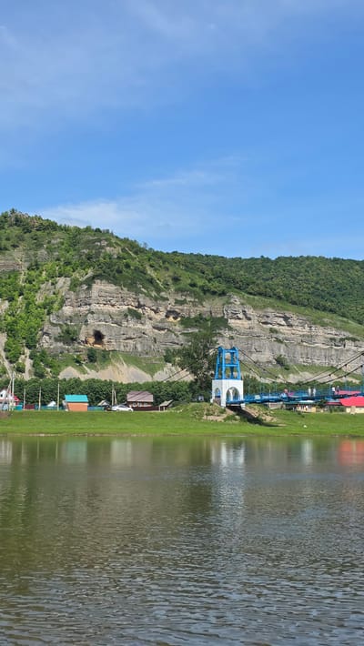A panoramic, eye-level, full shot captures a scenic view of the Tashtyk River in Tash-Asty, Russia. The river occupies the foreground, its surface rippled and reflecting the sky and surrounding landscape. On the far bank, a lush green field slopes gently upwards, dotted with small, colorful houses featuring red and blue roofs. A white and blue suspension bridge with pedestrian traffic crosses the river, its towers a focal point. Behind the village, a massive, layered rock formation rises dramatically, its sheer face covered in dense green foliage. The sky above is a vibrant blue with scattered white clouds, indicative of a clear, sunny day.