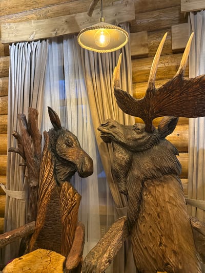 This media file captures a close-up view of two intricately carved wooden moose sculptures. The larger moose, positioned in the upper portion of the frame, is depicted from the head and impressive antlers down its body. Its textured, dark brown wood carving showcases detailed features of its face, including its eye and snout, with the large, branching antlers prominently displayed.

Below the larger moose is a smaller, similarly carved moose head, emerging from what appears to be a wooden branch structure. This smaller carving also exhibits detailed wood texture, emphasizing the animal's features.

The sculptures are set against a backdrop of light-colored, layered curtains, which are pulled to the side, revealing what might be a window or an opening. To the left, a vintage-style light fixture with a clear bulb and a rope-wrapped lampshade hangs, casting a warm glow. The background consists of a rustic wooden wall and ceiling, suggesting an interior setting, possibly a lodge or a cabin. The overall ambiance is warm and inviting, with the natural wood and the soft lighting creating a cozy atmosphere. The carvings themselves are the primary focus, highlighting the craftsmanship and the subject matter. No people are visible in the frame. The time of day appears to be evening or indoors with artificial lighting. There is no visible text in the image.