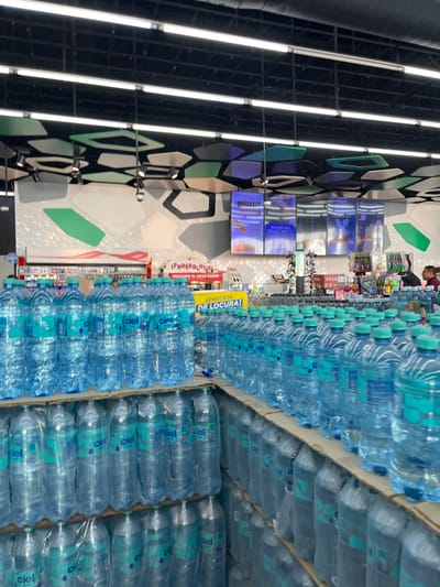The image displays an aisle inside a retail store, likely a supermarket or a large discount store, located in San Buenaventura, Mexico. The primary subject is a vast quantity of bottled water, predominantly from the "Ciel" brand, stacked high in bulk packages on multiple shelves or pallets that extend into the foreground and background.

In the mid-ground, a person wearing a red or brown shirt is visible near other merchandise, suggesting a customer browsing or a staff member working. Another figure is faintly seen further back. The store's interior features modern design elements, including a dark industrial-style ceiling with prominent linear fluorescent lights. One wall is adorned with decorative geometric panels in various shades of black, gray, green, and purple.

Several digital screens are mounted on the wall, displaying promotional content. Visible text on one screen reads "¡PRUÉBALO DESCUBRE EL GRAN SABOR!", encouraging customers to "Try it, discover the great flavor!" and possibly showing a coffee-related image. A bright yellow sign prominently displays "¡PRECIOS DE LOCURA!", translating to "Crazy Prices!", indicating a sale or special offer for the bottled water. Other small text elements like "POSA" and "201" are visible on shelf labels. The scene is brightly lit by artificial lights, typical of an indoor commercial environment.