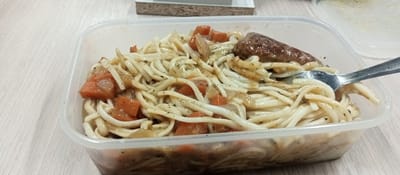 A close-up, high-angle shot shows a clear plastic food container filled with noodles, diced tomatoes, and a piece of sausage. A silver spoon is partially submerged in the food, resting on the rim of the container. The food appears to be in a light-colored sauce, seasoned with herbs. The container is situated on a light-colored, textured surface, possibly a wooden table. In the background, a blurred white object and a darker object are visible, suggesting an indoor setting. The overall lighting is neutral. There are no people visible in the image. The scene does not provide any specific information to connect it to Petropavl City Administration, Kazakhstan.