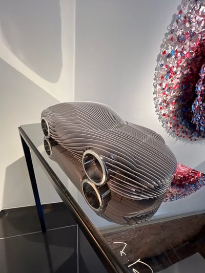 The image shows a sculpture of a car, made of thin, curved metal strips, displayed on a reflective surface. The sculpture is in a gallery or exhibition space. To the right, there is a circular artwork composed of small, colorful elements that appear to be butterflies. The background is a white wall with shadows cast by lighting fixtures, creating a sense of depth. The floor is dark and reflective. The overall impression is of a modern art installation.

This scene is located in the city of Honfleur, France. Specifically, it is inside the Studio of the artist Jean-Marc de Pas.