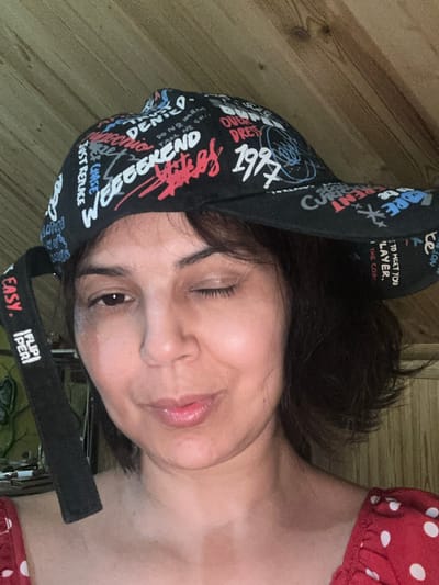 A woman is shown in a close-up, medium shot, wearing a black baseball cap with a graffiti-style print. She is looking at the camera and winking with her right eye. The cap has various words and phrases printed on it, including "WEEKEND," "DENIED," "1997," and "EASY." There is also a black strap hanging from the cap, with the words "FLIP PER" printed on it. The woman has dark, chin-length hair and is wearing a red top with white polka dots. The background appears to be a wooden wall. The image does not provide enough information to determine the location in the city of Kalininskaya, Russia.