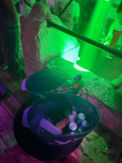 The image captures an outdoor scene at night, likely a bar or party venue in Canet-en-Roussillon, France. The lighting is dominated by vibrant green and pink neon lights, creating a club-like atmosphere. In the foreground, two large black buckets are filled with ice and beverages. The upper bucket contains several bottles, including one with a blue and red label, and a distinct yellow can with red lettering. The lower bucket is filled with numerous cans, predominantly red and silver, with one yellow can featuring red lettering clearly visible. In the background, blurred figures of people are visible, illuminated by the colorful lights, suggesting a social gathering or event. A rough, stone-like structure with a metal railing runs along the left side of the image, possibly a barrier or decorative element. The ground beneath the buckets appears to be wooden planks.