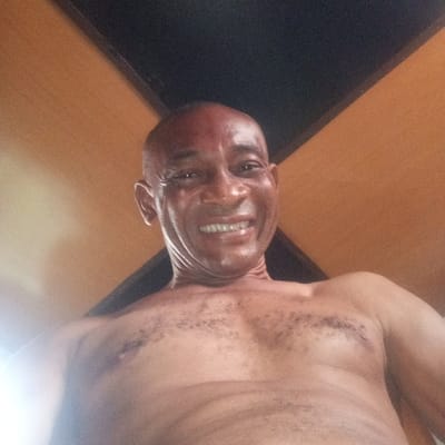 A shirtless adult male with dark skin and a bald or closely shaven head smiles broadly directly at the camera. His face appears glistening, and a slight redness is visible on his forehead. He exhibits some chest hair. The image is captured from a low angle, looking upwards at the subject. The background consists of a geometric pattern formed by intersecting dark and light orange-brown sections, suggestive of an interior wall or ceiling. The image does not contain details to identify a specific location within Koko, Nigeria.
