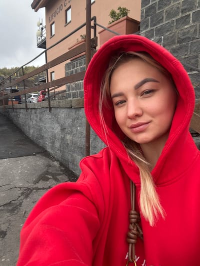 A young woman wearing a red hooded sweatshirt is posing for a photograph outside a building. A sign on the building reads "RIFUGIO G.S." The location is Nicolosi, Italy. The background consists of stone walls, a parking lot with cars, and the overcast sky.