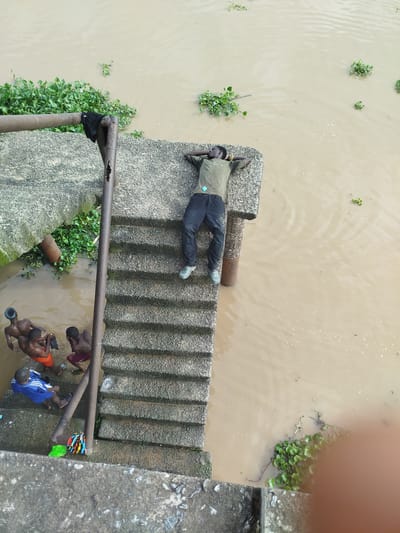 A high-angle medium shot shows a man lying on his back with his hands behind his head on a concrete ledge next to murky brown water in Yenegoa, Nigeria. The man is wearing a green t-shirt, black pants, and light blue shoes. He has a light blue device with wires attached to his shirt. Below the ledge, several young boys are in the water. One boy, wearing orange shorts, is being pulled up by another boy. Another boy, wearing maroon shorts, stands in the water. A boy in a blue shirt and white stripes sits on the concrete stairs. Colorful objects, including a green brush and striped fabric, are on the stairs. Water hyacinths float on the surface of the water. A rusty metal pole runs vertically along the stairs and attaches to a horizontal bar that extends across the top of the ledge.