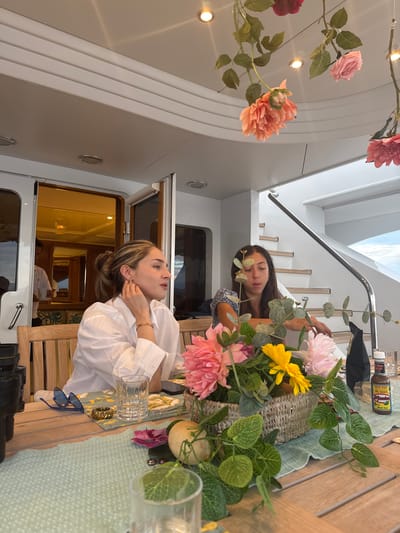 The image depicts two women seated at a wooden table aboard a boat, likely a yacht, in an unidentified location in Mexico. One woman, wearing a white shirt, is seated facing left, appearing to adjust an earring. The other woman, wearing a blue and white top, is seated facing away from the viewer, slightly behind the first woman. A floral arrangement in a woven basket serves as a centerpiece on the table, alongside glassware, sunglasses, and other unidentifiable objects. Overhead, artificial flowers hang from the ceiling. A staircase is visible in the background, leading to another level of the boat. Through a doorway behind the woman in the white shirt, a glimpse of the boat's interior, including what appears to be a steward, can be seen. Portholes are visible on the walls of the boat, and the overall environment suggests a luxurious setting.