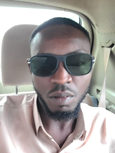 A man is taking a selfie in a car. He is wearing sunglasses and a light-colored collared shirt. The background shows the interior of the car, including a headrest and a seatbelt. The lighting suggests it is daytime. The image is taken from a close-up perspective, focusing on the man's face and upper body. The context of Karu, Nigeria is not depicted within the visual information of the image.