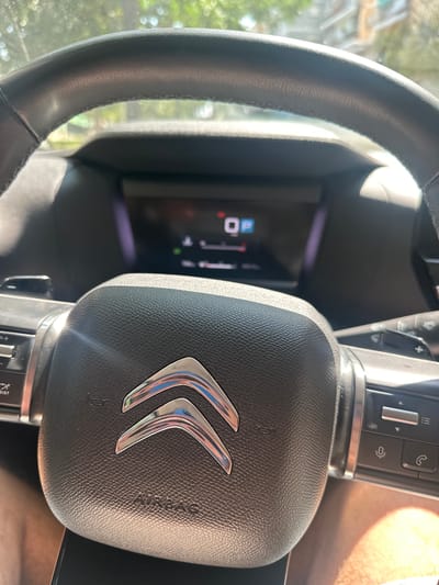 The media file is a close-up, eye-level shot of the interior of a car, specifically focusing on the steering wheel and dashboard. The steering wheel is black and appears to be made of leather, with stitching visible along its rim. In the center of the steering wheel is a silver emblem consisting of two chevrons, the logo of the car manufacturer, Citroën. The word "AIRBAG" is embossed on the steering wheel's central pad.
To the left of the steering wheel, a portion of the car's digital dashboard is visible. It displays a digital gauge with a reading of "0" and a lock symbol, along with other indicators that are partially obscured. On the right side of the steering wheel column, there are several control buttons, including what appear to be volume controls and buttons with icons for a phone and a cruise control or speed limiter system.
The scene is brightly lit, suggesting it is daytime. Sunlight is casting reflections on the steering wheel and dashboard, and a warm, hazy light is visible in the upper right portion of the frame, possibly from sunlight entering through the windshield or window. The background is blurred, showing hints of green foliage and structures outside the car, indicating it is parked outdoors. The overall impression is a snapshot of a car's interior on a sunny day in what is identified as Benimàmet-beniferri, Spain. No people are visible in the image.
