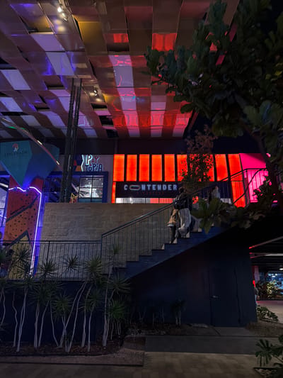 The image captures a nighttime scene in San Pedro Cholula, Mexico, featuring an exterior view of a complex containing several businesses and recreational areas. The building has a modern design with a mix of materials, including a lattice-like wall on the left illuminated with alternating red and purple lights. A prominent red sign with the word "CONTENDER" vertically displayed identifies a business. Stairs lead up to an entrance above. Below, a sign reading "PLAYTOPIA" is visible, and a brightly lit climbing wall with blue and orange sections is positioned at the bottom. The surrounding environment includes greenery along the stairs and a sidewalk leading into the frame on the right.