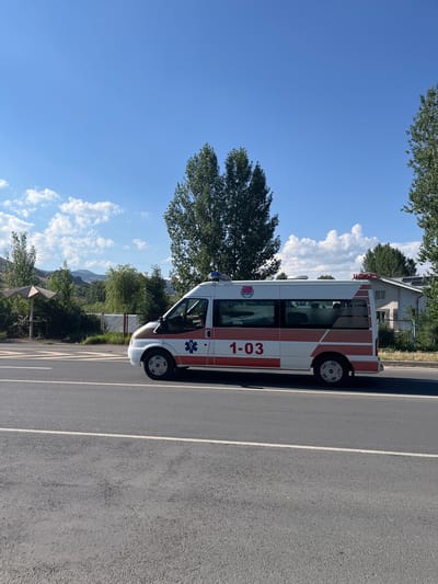 The image shows an ambulance parked on an asphalt road. The ambulance is white with red stripes and has the number "1-03" written on its side. A red cross symbol is also visible on the vehicle. The scene appears to be outdoors, with a clear blue sky and some white clouds. Trees and buildings are visible in the background. The image is oriented vertically, so the ambulance and surroundings are sideways. The time of day appears to be daytime, likely sunny, given the bright lighting and blue sky. The setting seems to be a rural or suburban area in Gladzor, Armenia, as suggested by the presence of mountains in the distance and the style of the buildings. There is no discernible activity taking place; the ambulance is stationary. The overall impression is a bright and clear day. Text visible on the ambulance includes "TRANSIT" and "1-03". A red sign with some text is also visible on the building in the background, but the text is not legible.