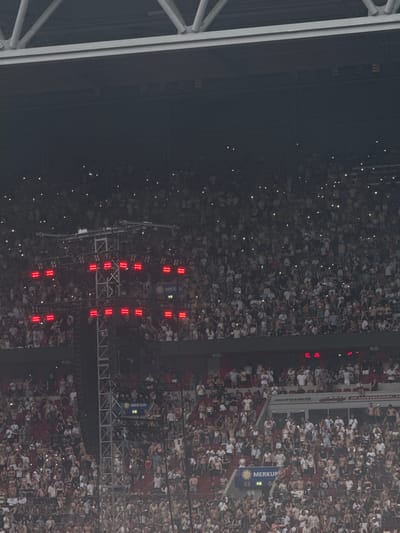 This aerial, high-angle shot depicts a large crowd of people packed into a stadium or arena at night, illuminated by numerous small, bright lights held by individuals in the audience. The scene suggests a concert, sporting event, or other large gathering. The audience members are mostly seen from above, with many standing and facing towards a central area or stage, indicated by the presence of lighting rigs and what appears to be a stage structure.

The lighting rigs consist of vertical arrays of red lights, forming an abstract pattern or possibly displaying text or symbols, though the specific details are difficult to discern. The crowd is dense, with many individuals wearing light-colored clothing, which stands out against the darker surroundings and the dimmer lighting. The overall atmosphere seems to be one of excitement and engagement, with the audience actively participating by holding up lights.

Visible text on a banner reads "MERKUR" with a sun logo and the numbers "03" and "04" below it, suggesting a sponsor or part of an advertisement within the venue. Another partial sign reads "IN DÜSSELDORF". The presence of "Düsseldorf" confirms the location. The time of day is clearly night, and the weather is not discernible from the image. The composition is dominated by the mass of people and the artificial lighting from both the stage and the audience.