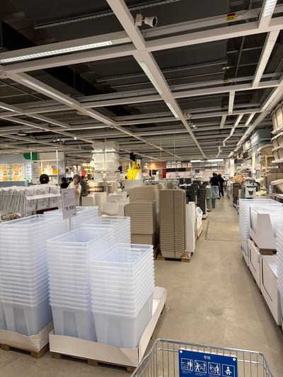 This is an indoor shot of a retail store, likely IKEA, located in Goyang-si, South Korea, based on the Korean signage visible. The scene depicts a wide aisle in a home goods store, with numerous storage containers, shelves, and display racks. Fluorescent lights are mounted on a grid-like ceiling structure. In the foreground, large, clear plastic storage bins are stacked on pallets. To the left, more rows of storage containers are visible, along with shelving units displaying various items. In the middle ground, a woman with curly hair is looking upwards, seemingly at a display or product. Further down the aisle, two individuals are walking away from the camera, and more shelves and storage solutions are apparent. A sign with Korean text is visible, and another sign in the foreground reads "₩9,900", indicating pricing. The floor is concrete. The overall atmosphere is that of a busy retail environment focused on home organization products.