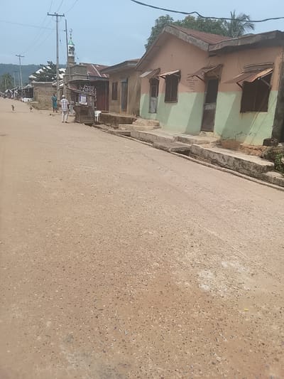 The image depicts a street scene with buildings, people, and a mosque in the background. The street is paved with a light-colored, coarse material. On the right side of the street, there are buildings with faded paint, some with corrugated metal awnings over the windows. There is a drainage channel along the side of the road. On the left side, there are more buildings and a few people walking. A mosque with a green-domed minaret is visible further down the street. Power lines are strung overhead. The overall impression is of a developing or rural area, not a major city.
This scene is not located in Birmingham, United Kingdom. There are no recognizable landmarks, architecture, or environmental cues that would suggest this location is Birmingham. The style of buildings and the general appearance are inconsistent with urban environments in the United Kingdom.