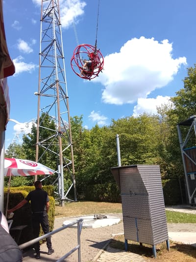 This media file captures a sunny day at an amusement park in Rammingen, Germany. The central focus is a towering metal structure, likely a thrill ride, with a red spherical cage suspended from the top. Inside the cage, two people are visible, appearing to be enjoying the ride as they are swung high into the air. The sky above is a vibrant blue, dotted with fluffy white clouds, suggesting pleasant weather.
In the foreground, a man in a black polo shirt and pants stands near a red and white umbrella, seemingly an employee or attendant. To his right, a metallic enclosure with a diamond plate pattern stands on blue legs, possibly a control booth or equipment housing. Behind the man and the umbrella, lush green trees form a dense backdrop, and further in the distance, the silhouette of a Ferris wheel can be seen, hinting at the park's larger attractions. The ground is paved with interlocking bricks and patches of grass. The overall scene conveys a sense of fun and excitement, typical of a day at an amusement park. The time of day appears to be midday, given the bright sunlight and strong shadows. No visible text is discernible in the image.
