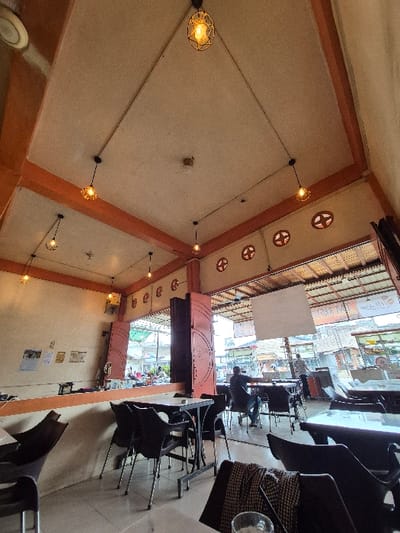 The image displays the interior of an open-air cafe or restaurant, looking out onto a street in the city of Manetunong, Indonesia. The establishment features a light beige ceiling with prominent orange structural beams. Multiple pendant lights with decorative cage-like shades hang from black cords, illuminating the space. Circular, cross-patterned ventilation cutouts are visible along the upper walls. The floor is covered with light-colored tiles.
Throughout the interior, black square tables with metal bases are paired with dark plastic or metal chairs. In the immediate foreground, a table holds a folded plaid garment and a clear glass. Towards the middle-right, a male individual with dark skin, wearing a dark shirt and blue jeans, is seated at a table, facing away from the camera. Another person is visible standing near the entrance, further in the background.
The open entrance, framed by dark reddish-brown folding doors, provides a view of a busy street scene. Several motorbikes are visible on the street, along with pedestrians, other buildings, and various commercial signs, one partially readable as "RESTORAN." A large white rectangular screen or sign is suspended above the outdoor area. A partial view of a ceiling fan is seen in the upper left, and an overhead speaker is mounted on a beam on the left.