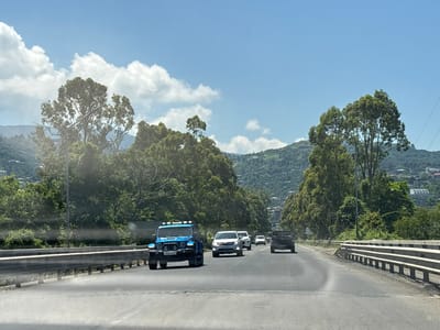 A blue Jeep Wrangler is traveling on a highway in a mountainous, tree-lined area, possibly near Akhalsopeli, Georgia. The Jeep is in the left lane, facing away from the camera, with its headlights on. Several other vehicles are on the highway, including a white SUV, a black pickup truck, and two other white cars. The road is asphalt and has lane markings. Guardrails are visible on both sides of the road. The sky is blue with scattered white clouds. Lush green trees and vegetation cover the surrounding hills, with some buildings visible on the slopes in the distance. The day is bright and sunny.