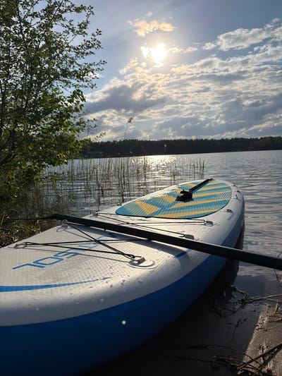 The image depicts a paddleboard resting on the edge of a body of water, likely a lake or river, in Selychka, Russia. The paddleboard is partially in the water and partially on the shore. A paddle is placed across the board. The water reflects the sky, which features scattered clouds and the sun. The shoreline is visible with vegetation.